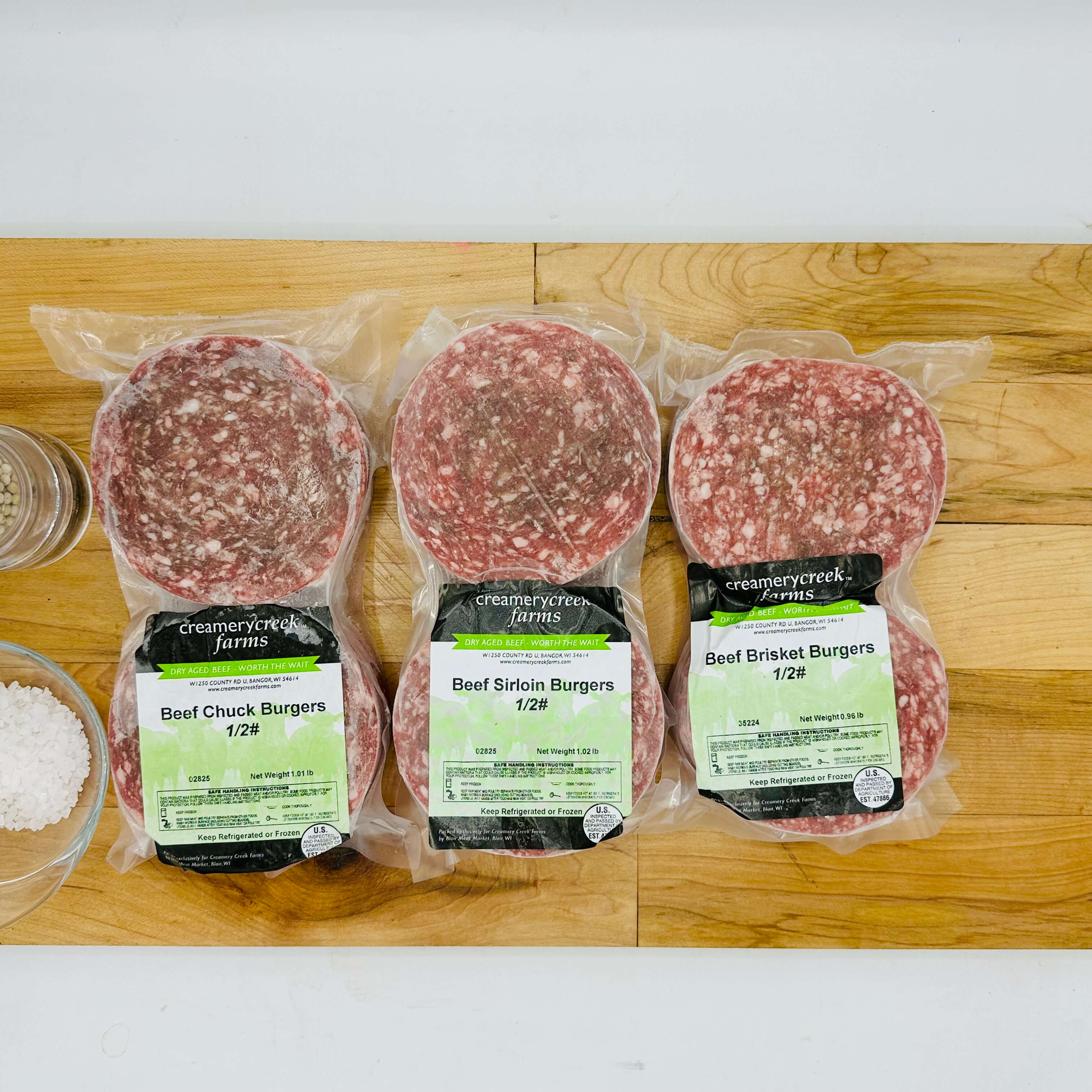 Three packages of ground beef labeled 'Beef Chuck Burgers', 'Beef Sirloin Burgers', and 'Beef Brisket Burgers' on a wooden surface.