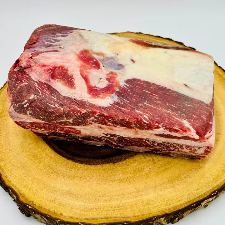 A raw cut of beef with marbling, showing red muscle tissue and white fat, resting on a round wooden slice against a light gray background.