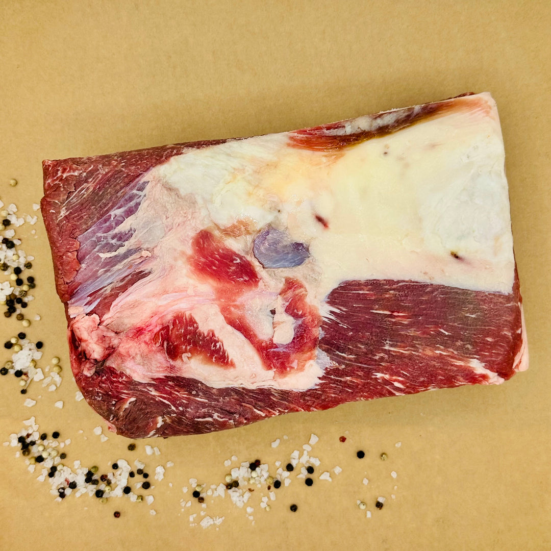 A raw cut of marbled beef with fat, photographed from above on a tan background with scattered coarse salt and peppercorns.