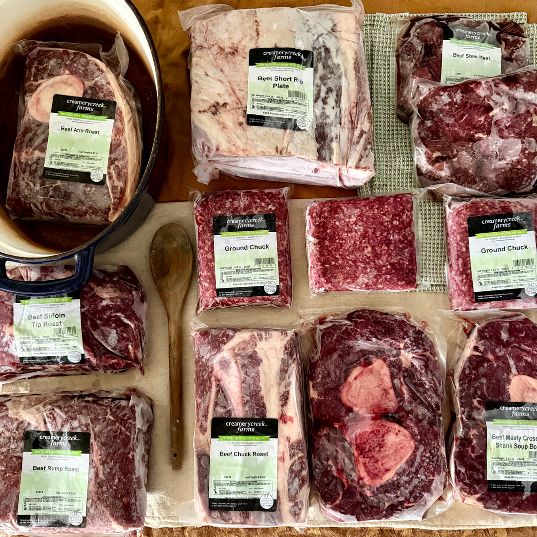 An assortment of packaged beef cuts from Creamery Creek Farms arranged on a wooden surface, including beef short rib, ground chuck, beef chuck roast, beef pot roast, and beef brisket, with a wooden spoon and a dark pot visible.