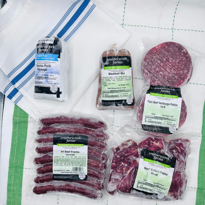 Five packages of Creamery Creek Farms meat products arranged on a white quilted blanket with green stripes, including Shrimp Pork Sauté, Meatball Mix, Pan Beef Hamburger Pattes, All Beef Franks, and Beef Brisket Frites.