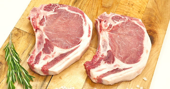 Two raw pork chops on a wooden cutting board with rosemary and salt.