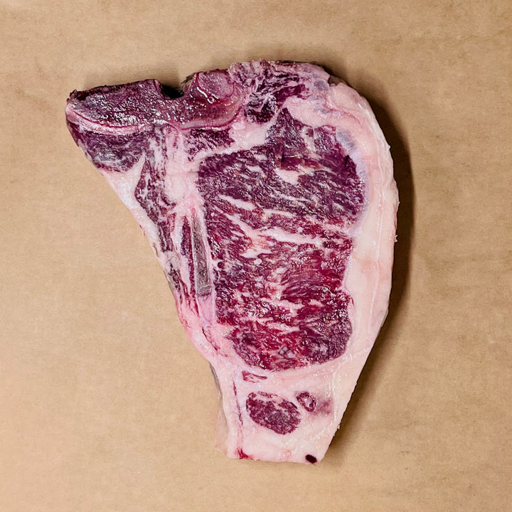A raw, highly marbled t-bone steak with a bone, displayed flat on a textured brown paper background.
