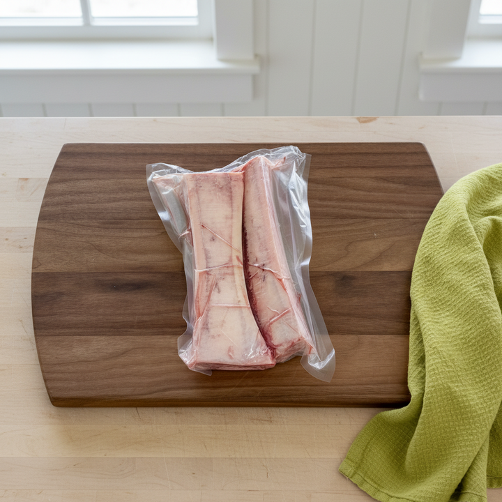 Two vacuum-sealed bone-in marrow beef bones, resting on a dark wood cutting board with a green cloth on a light wood table.
