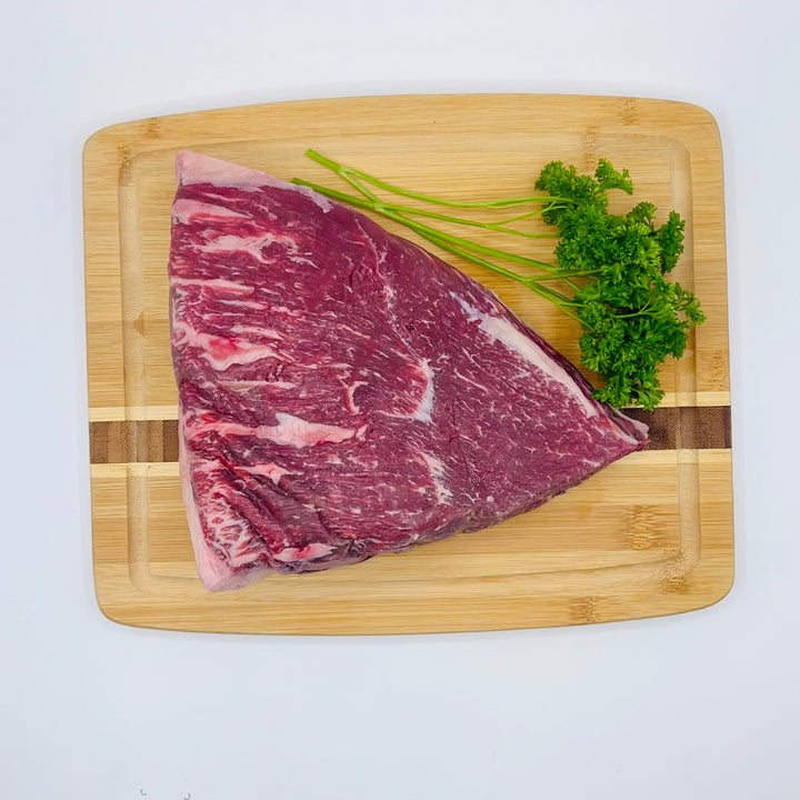 A large raw cut of marbled red beef picanha rests on a rectangular wooden cutting board, with a small bunch of green parsley placed on top, photographed against a plain white background.