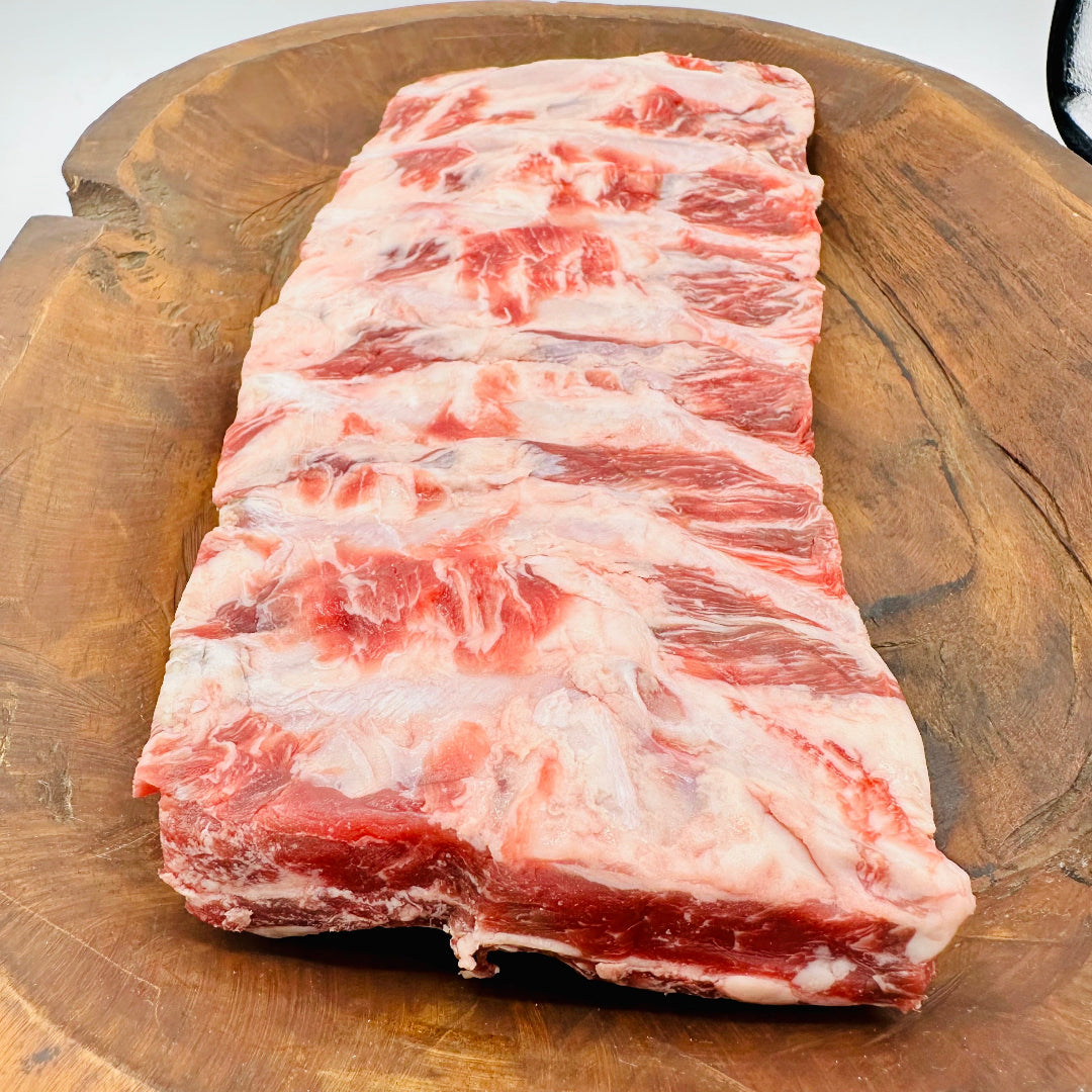 A raw rack of beef back ribs with visible marbling sits on a large round wooden cutting board against a light neutral background.