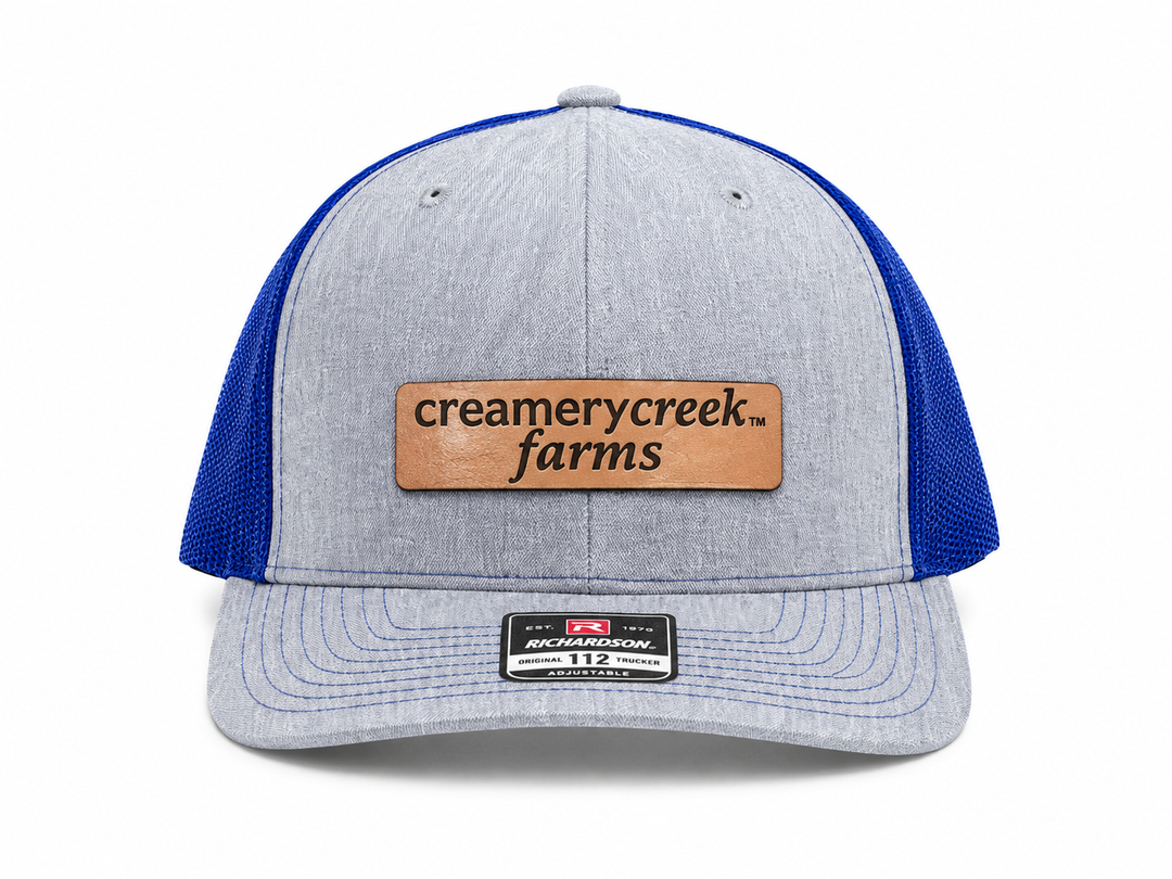 A light heather gray and royal blue trucker hat with a tan leather patch on the front that reads 'creamy creek farms'.