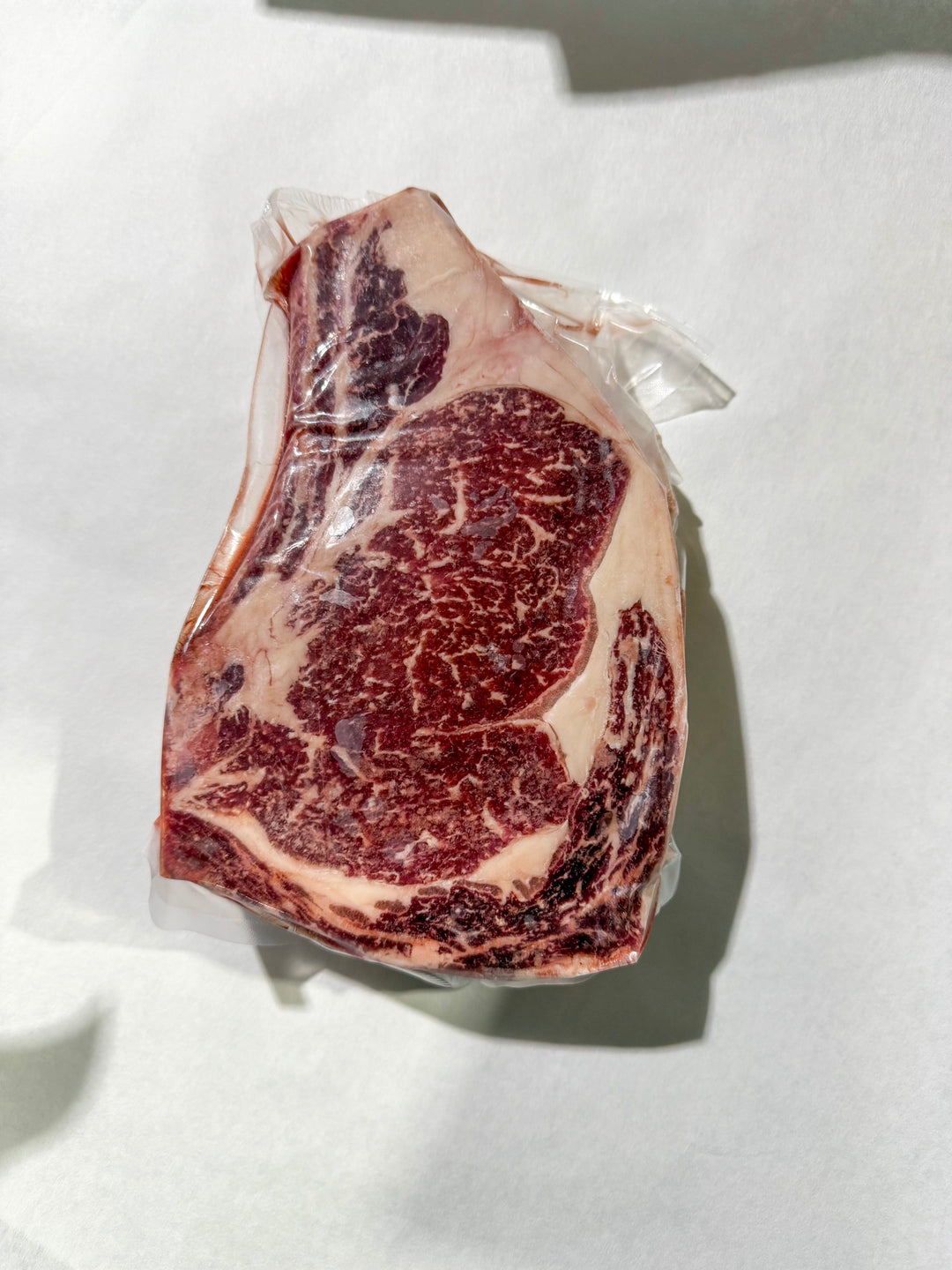 A raw marbled bone in ribeeye steak in clear plastic packaging, photographed from above on a white surface with a hard shadow.
