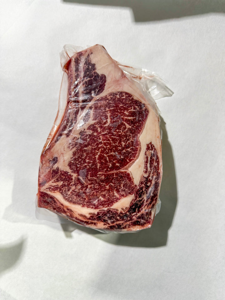 A raw marbled bone in ribeeye steak in clear plastic packaging, photographed from above on a white surface with a hard shadow.
