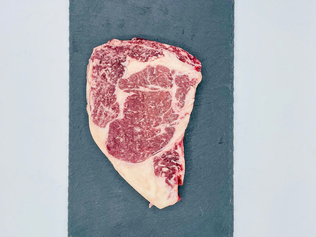 A raw marbled steak with red meat and white fat, placed on a dark slate serving board against a light gray background.