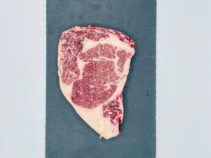 A raw marbled steak with red meat and white fat, placed on a dark slate serving board against a light gray background.