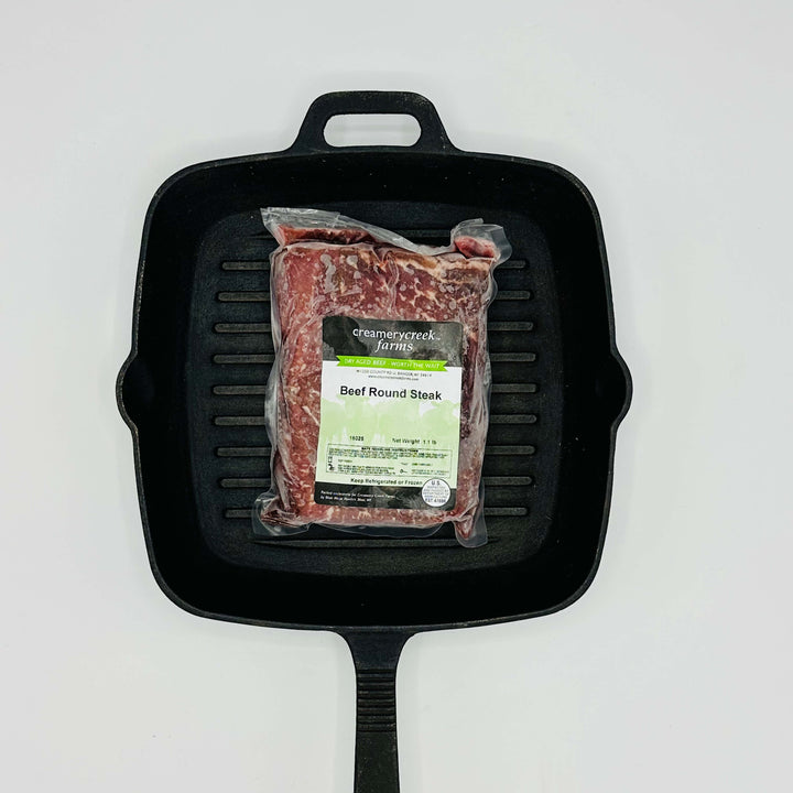 Packaged beef round steak on a black cast iron skillet with a white background