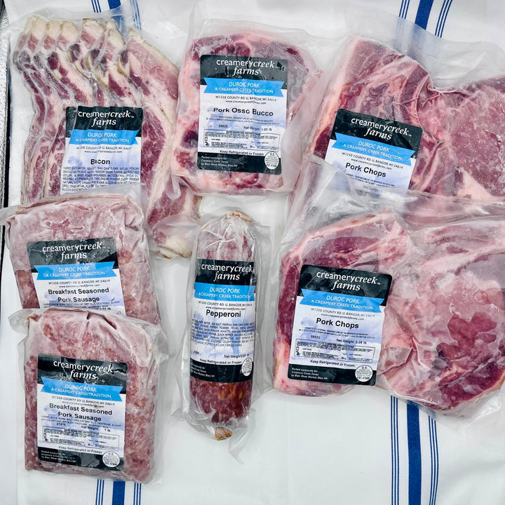 A variety of packaged pork from Creamery Creek Farms arranged on a white surface with blue stripes. The products include bone-in Pork Chops, Pepperoni, osso bucco, and Pork Sausages