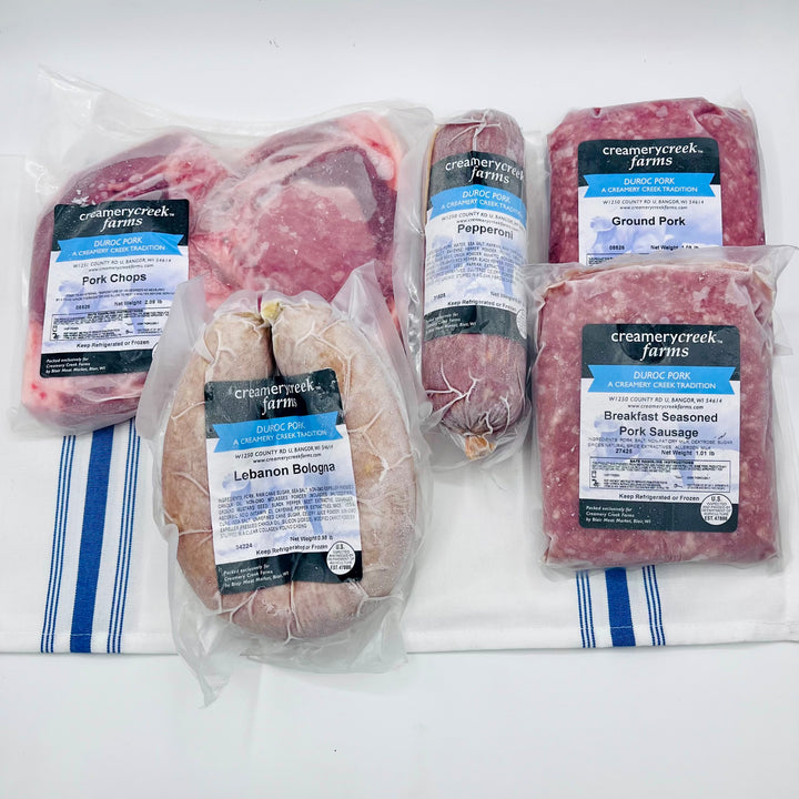 A variety pack of Creamery Creek Farms cured meats and ground pork, including Pork Chops, Lebanon Bologna, Pepperoni, Ground Pork, and Breakfast Seasoned Pork Sausage, all in clear plastic packaging with blue and white labels, arranged on a white cloth with blue stripes.