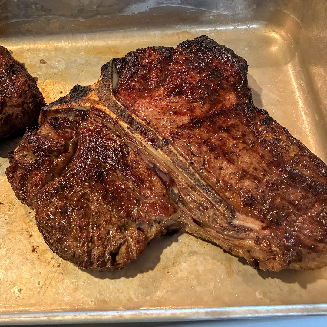 A close-up of a cooked porterhouse steak with a dark brown and charred exterior, resting in a metal baking pan.