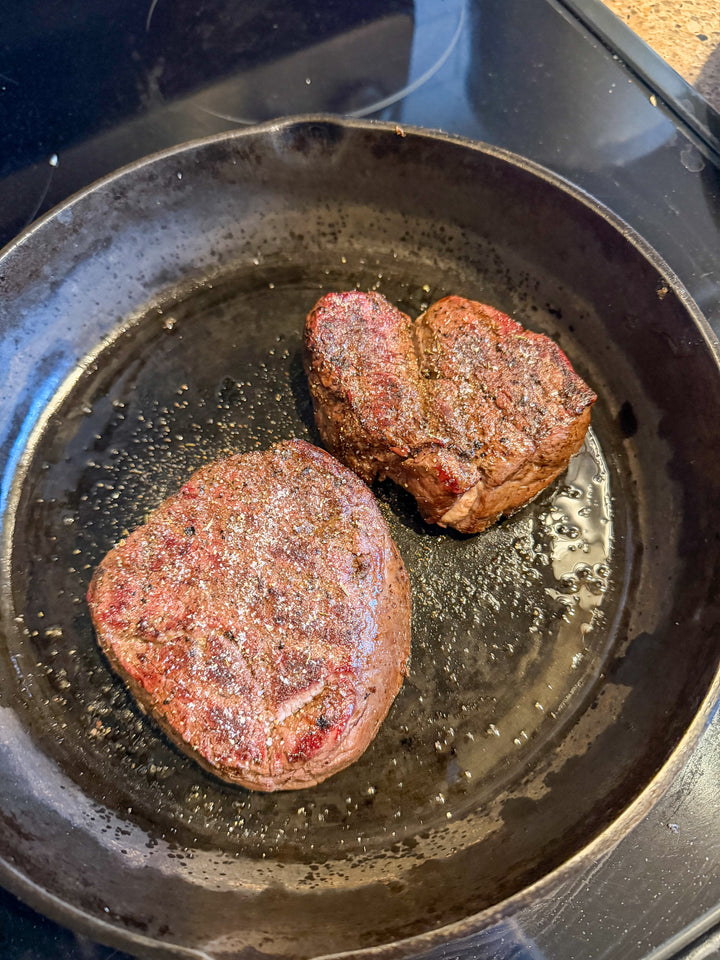 Two seasoned steak beef filets cooking in a dark cast iron skillet on a black stovetop.