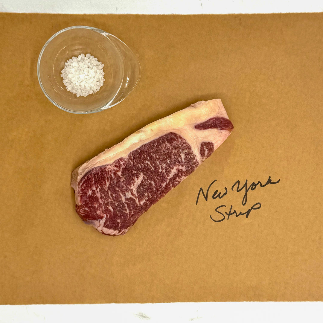 A raw marbled beef New York Strip steakn, placed on a brown paper background with a small clear bowl of coarse salt and handwritten text reading 'New York Strip'.
