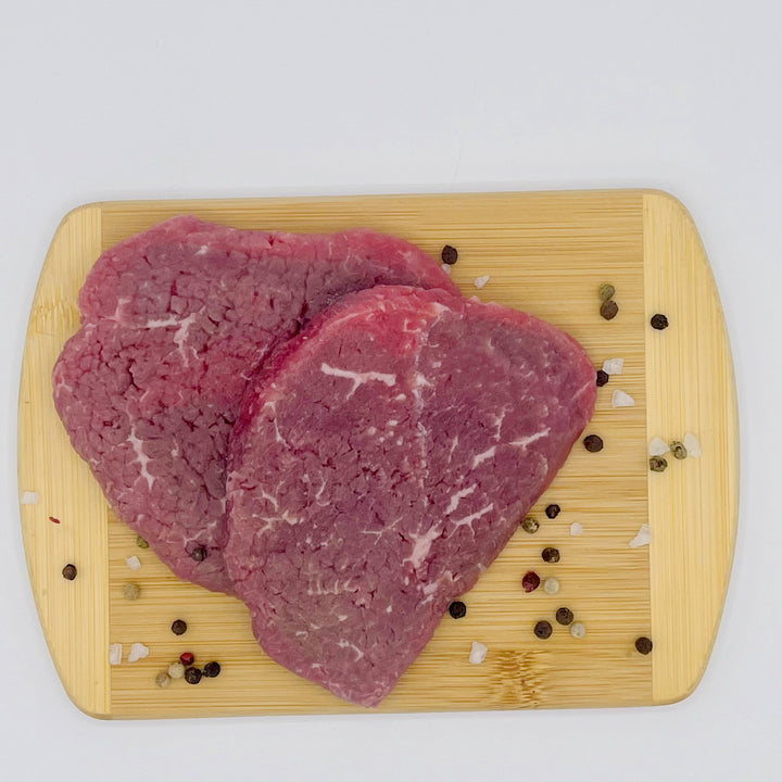 Two raw beef cube steeaks with visible marbling, arranged on a rectangular light wood cutting board with scattered coarse salt and peppercorns on a white background.