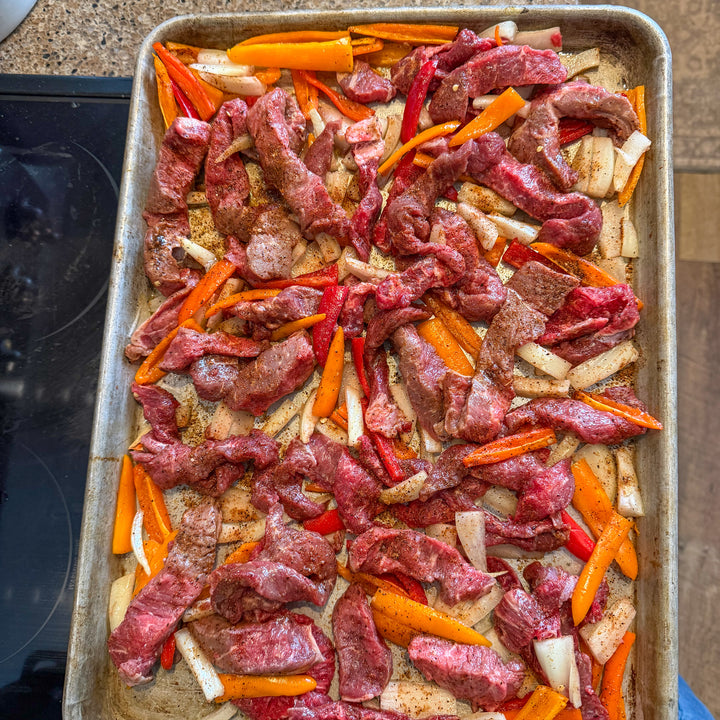 Raw sliced beef strips mixed with sliced carrots, red bell peppers, and onions on a metal baking sheet, photographed from above in a kitchen setting.