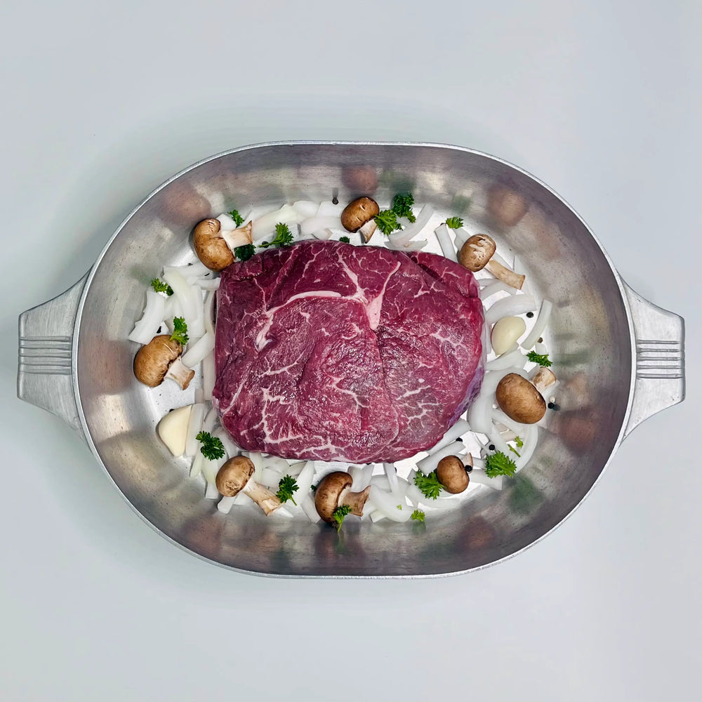 A raw beef roast with visible marbling, arranged in a round stainless steel roasting pan with sliced white onions, brown mushrooms, and small green herb pieces on a light gray background.