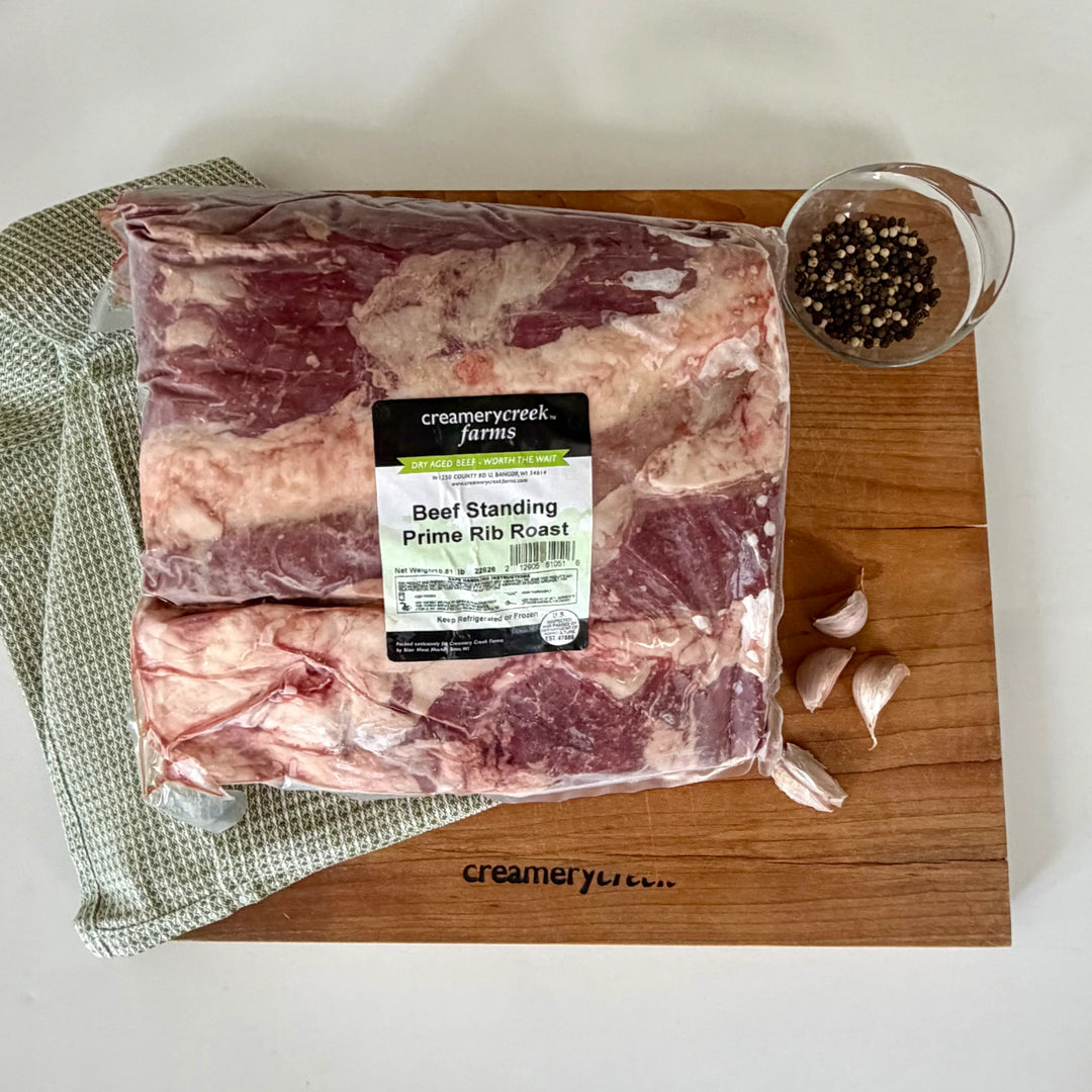 Packaged beef standing rib roast on a wooden cutting board with garlic cloves and a small bowl of peppercorns, branded Creamery Creek Farms.
