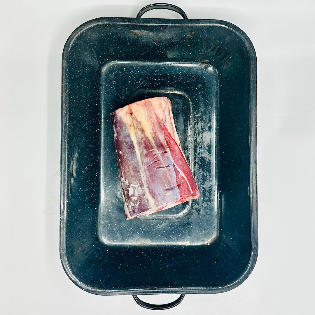 A raw cut of whole beef foreshank with fat marbling sits inside a dark rectangular roasting pan on a plain white background.