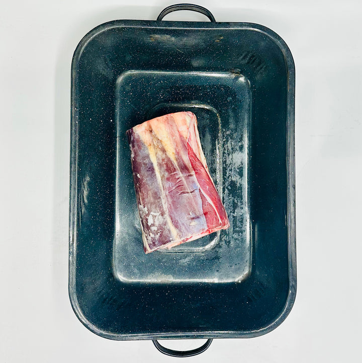 A raw cut of whole beef foreshank with fat marbling sits inside a dark rectangular roasting pan on a plain white background.